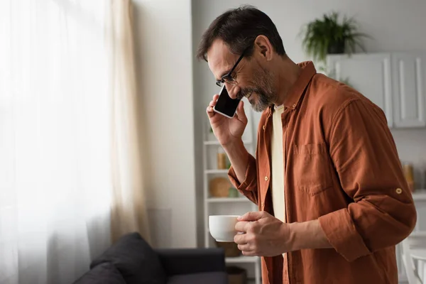 Vista laterale dell'uomo sorridente negli occhiali che tiene il caffè del mattino durante la conversazione sullo smartphone in cucina — Foto stock