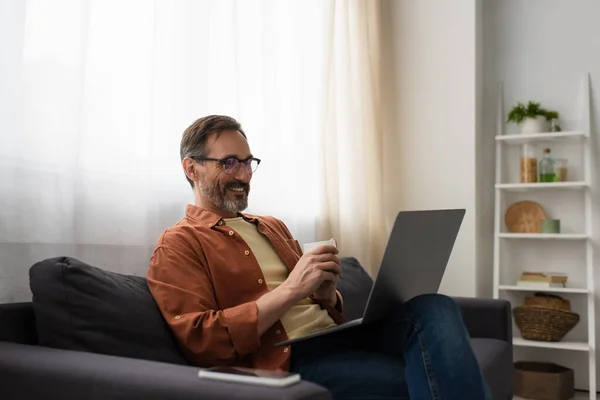 Homme joyeux dans les lunettes assis sur le canapé avec tasse de café et ordinateur portable — Stock Photo