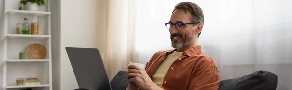 Homme gai dans des lunettes tenant tasse de café et regardant ordinateur portable, bannière — Photo de stock