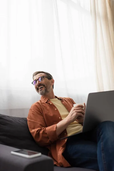 Joyful man in eyeglasses looking away while sitting on couch with laptop and coffee cup — Stock Photo