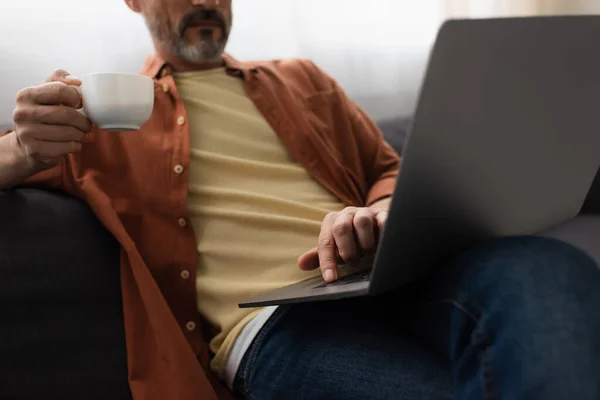 Cropped view of man sitting on sofa with coffee cup and using laptop — Stock Photo