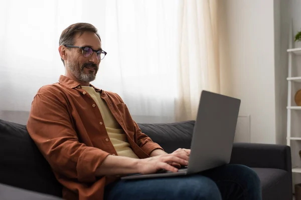 Homme barbu positif dans les lunettes assis sur le canapé et dactylographier sur ordinateur portable — Photo de stock