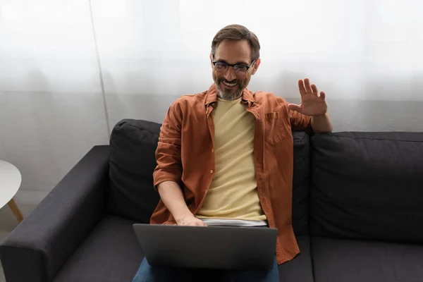 Щасливий бородатий чоловік в окулярах сидить на дивані і махає рукою під час відеодзвінка на ноутбук — стокове фото