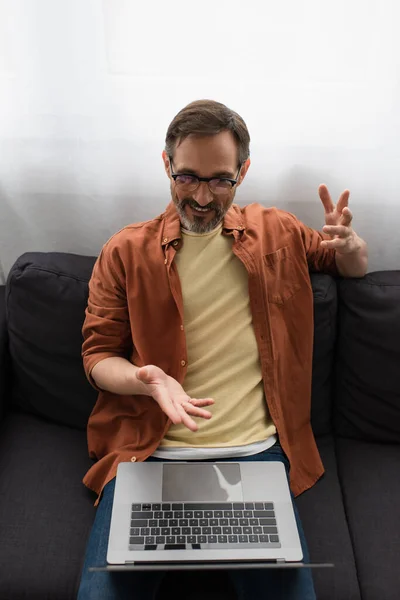 High angle view of smiling man in eyeglasses pointing at laptop during video chat on couch at home — Stock Photo