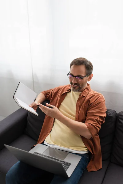 Lächelnder Mann mit Brille zeigt bei Videoanruf auf Laptop zu Hause auf leeres Notizbuch — Stockfoto