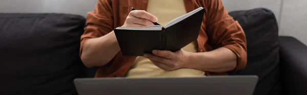 Vista recortada del hombre sentado en el sofá con el ordenador portátil borroso y la escritura en el cuaderno, pancarta - foto de stock