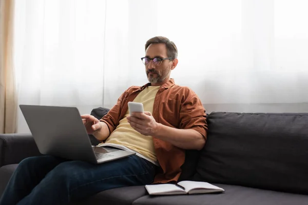 Mann mit Brille hält Smartphone in der Hand und zeigt auf Laptop, während er auf Couch neben leerem Notizbuch sitzt — Stockfoto