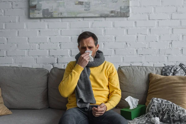 Sick man in warm scarf sitting on sofa at home and sneezing in paper napkin — Stock Photo
