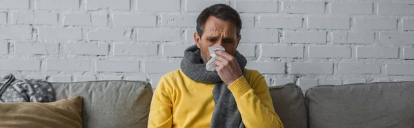 Homme malade dans une écharpe chaude souffrant de nez qui coule et éternuant dans une serviette en papier, bannière — Photo de stock