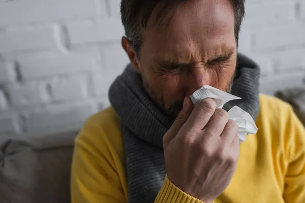 Malade dans un foulard chaud tenant une serviette en papier et éternuant les yeux fermés — Photo de stock