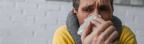 Malade en écharpe chaude souffrant de nez qui coule et tenant une serviette en papier près du visage, bannière — Photo de stock