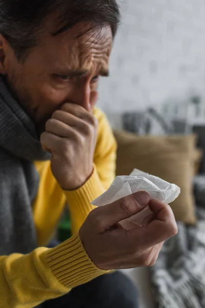Hombre enfermo en bufanda caliente sosteniendo servilleta de papel y estornudando en casa - foto de stock