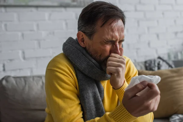 Hombre enfermo en bufanda caliente estornudando con los ojos cerrados mientras sostiene la servilleta de papel - foto de stock