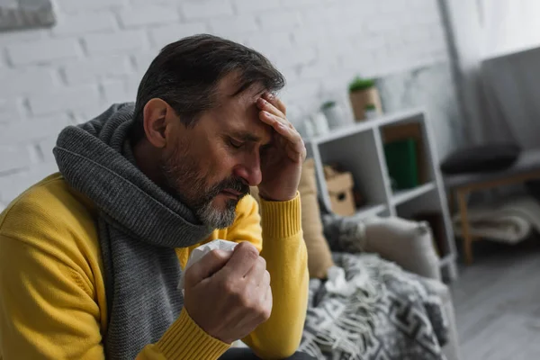 Хворий чоловік в теплому шарфі тримає паперову серветку і страждає від головного болю — стокове фото