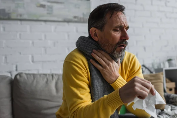 Хворий чоловік в теплому шарфі торкається болю в горлі і кашлю, тримаючи паперову серветку — стокове фото