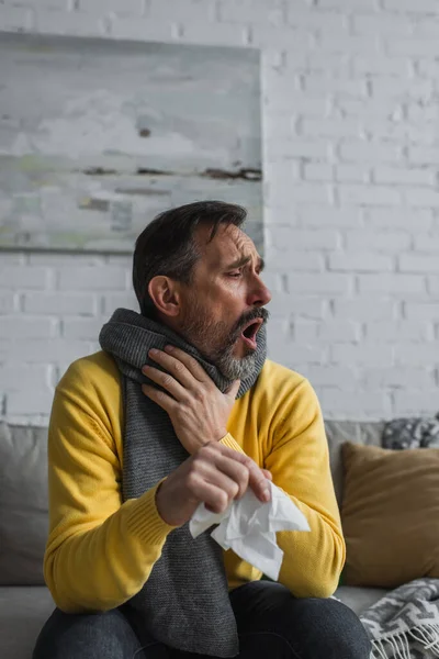 Uomo malato in sciarpa calda tenendo tovagliolo di carta e tosse sul divano — Foto stock