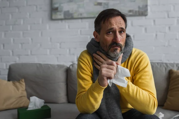 Hombre enfermo en bufanda caliente tocando dolor de garganta mientras sostiene la servilleta de papel y mirando a la cámara - foto de stock