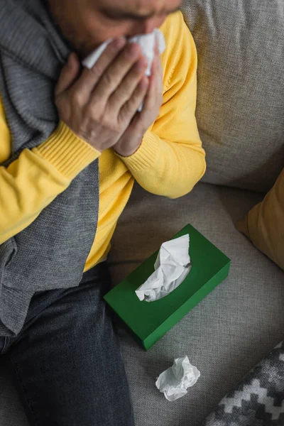 Erkrankter Mann niest, während er auf Couch neben Packung Papierservietten sitzt — Stockfoto