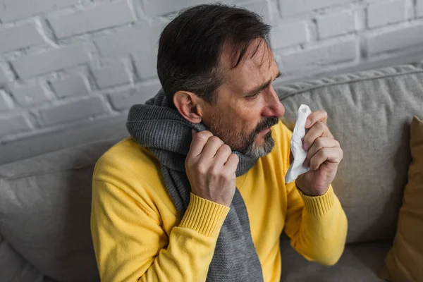 Kranker Mann in warmem Schal hält Papierserviette, während er zu Hause auf Couch sitzt — Stockfoto