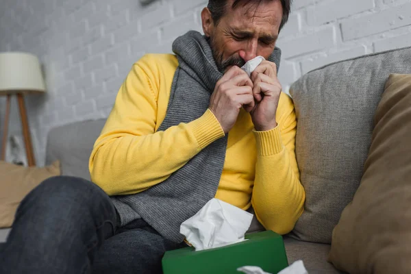 Sick man in warm scarf sneezing in paper napkin with closed eyes on couch — Stock Photo