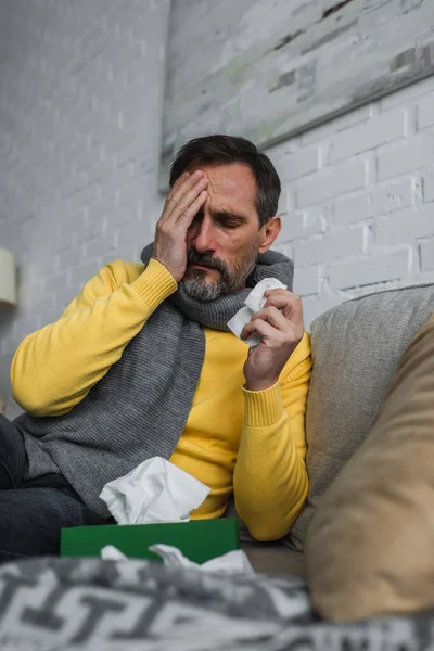 Больной человек затемняет лицо рукой, сидя на диване с бумажной салфеткой и страдая от головной боли — стоковое фото