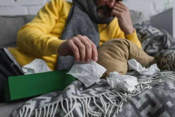 Partial view of sick man lying on couch with warm blanket and holding paper napkin — Stock Photo