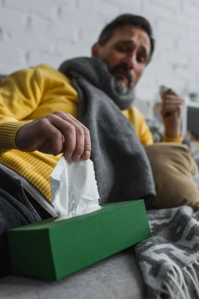 Hombre enfermo en bufanda caliente tomando servilleta de papel del paquete sobre fondo borroso - foto de stock