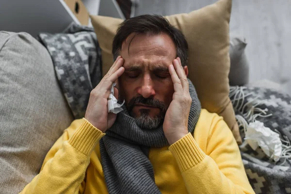 Top view of sick man in warm scarf touching head while lying on couch and suffering from migraine — Stock Photo