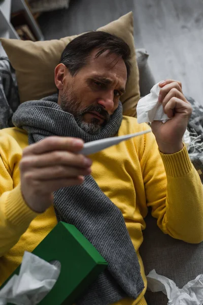 Хворий чоловік в теплому шарфі тримає паперову серветку і дивиться на електронний термометр — стокове фото
