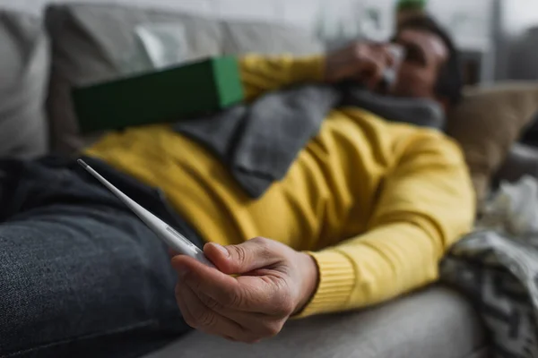 Selektiver Fokus des elektronischen Thermometers in der Hand eines kranken Mannes, der zu Hause auf dem Sofa liegt — Stockfoto