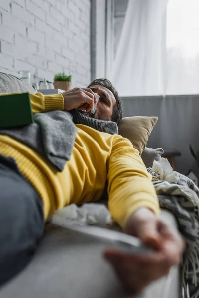 Diseased man sneezing in paper napkin while lying on couch on blurred foreground — Stock Photo