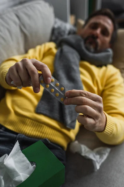 Kranker Mann in warmem Schal hält Tabletten in der Hand, während er auf Couch vor verschwommenem Hintergrund liegt — Stockfoto