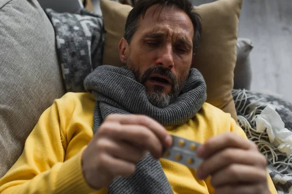 Homem doente em cachecol quente segurando pílulas borradas enquanto deitado no sofá — Fotografia de Stock