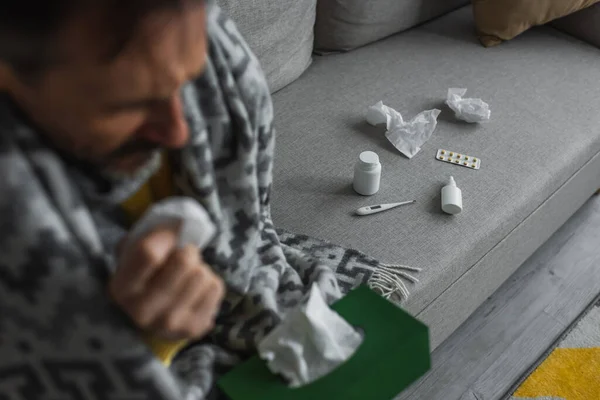 Homme malade avec des serviettes en papier assis sur le canapé près du thermomètre électronique et des conteneurs avec traitement — Photo de stock