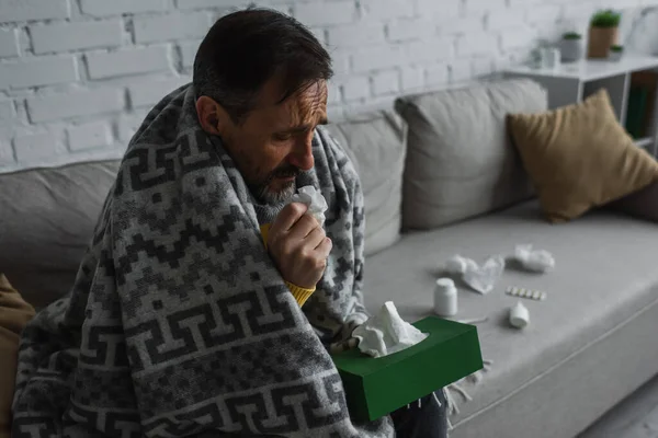 Хворий чоловік тримає пакет паперових серветок, сидячи на дивані біля контейнерів з лікуванням — стокове фото