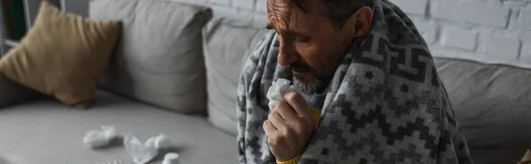 Больной человек сидит на диване под теплым одеялом и держит бумажную салфетку, баннер — стоковое фото