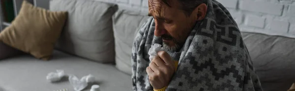 Sick man sitting on couch under warm blanket and holding paper napkin, banner — Stock Photo