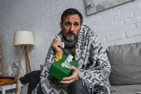 Hombre enfermo sentado en el sofá debajo de una manta caliente y sosteniendo el paquete de servilletas de papel - foto de stock