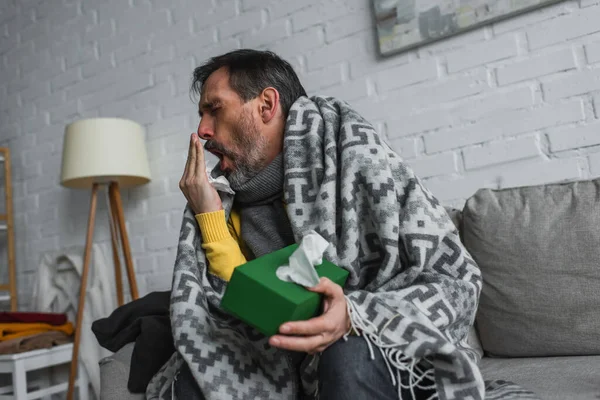 Чоловік вкритий теплою ковдрою, тримає пакет паперових серветок і кашляє на дивані — стокове фото