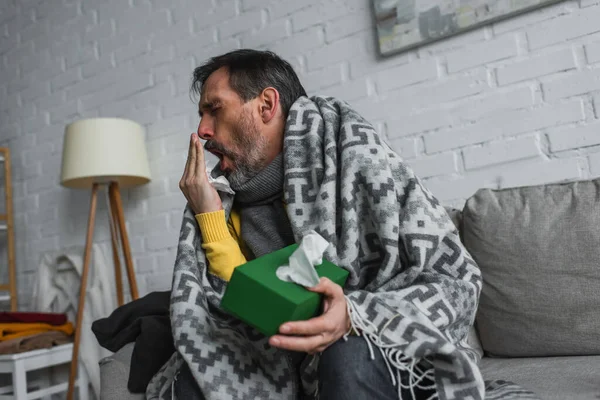 Man covered with warm blanket holding pack of paper napkins and coughing on sofa — Stock Photo