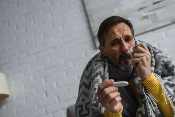 Sick man sitting under warm blanket and looking at electronic thermometer — Stock Photo