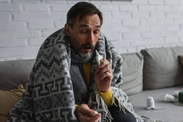 Hombre enfermo con los ojos cerrados sentado debajo de la manta caliente y la celebración de aerosol nasal - foto de stock