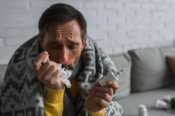 Uomo malato con gli occhi chiusi che soffre di naso che cola e tiene spray nasale con tovagliolo di carta — Foto stock