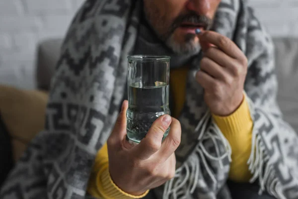 Vista ritagliata di uomo malato sotto coperta calda tenendo bicchiere d'acqua e prendendo pillola su sfondo sfocato — Foto stock