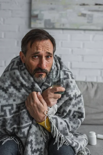 Hombre enfermo sentado debajo de una manta caliente con un vaso de agua y mirando a la cámara - foto de stock