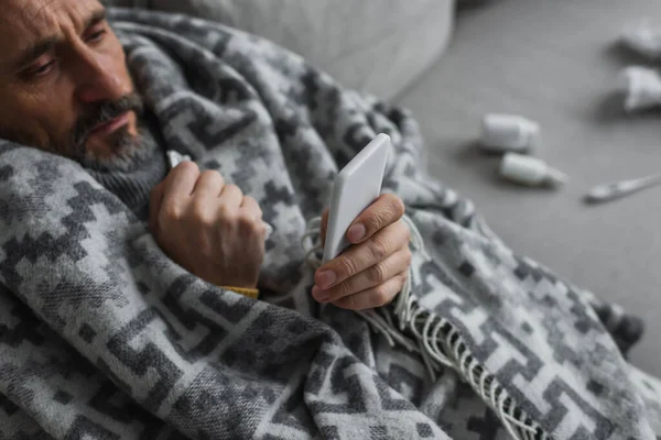Vista de ángulo alto del hombre enfermo envuelto en una manta caliente mirando el teléfono móvil - foto de stock