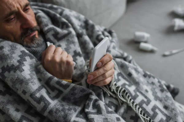 Vista ad alto angolo di uomo malato avvolto in una coperta calda guardando il telefono cellulare — Foto stock