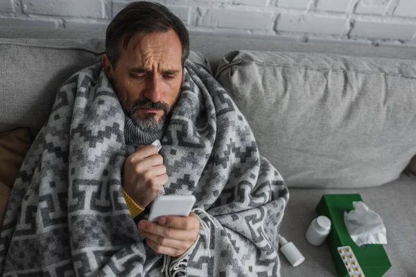 Man covered with warm blanket looking at smartphone while sitting near containers with treatment and pack of paper napkins — Stock Photo