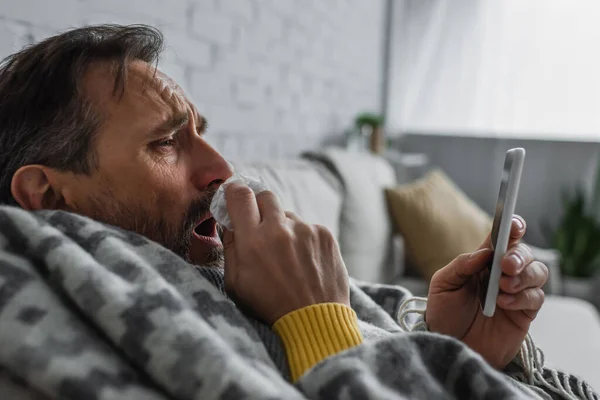 Sick man with paper napkin coughing while looking at mobile phone — Stock Photo