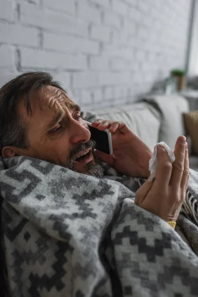 Больной человек завернутый в теплое одеяло держит бумажную салфетку и разговаривает по мобильному телефону — стоковое фото